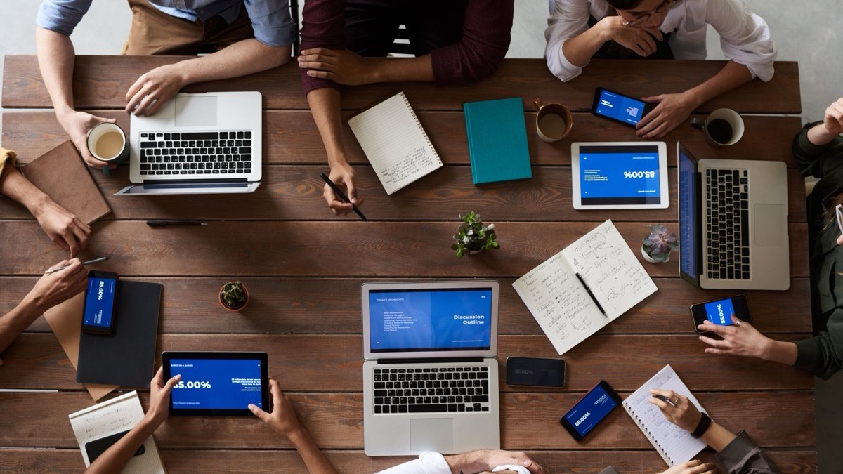A team around a wooden table with laptops, tablets and notebooks working through a project together