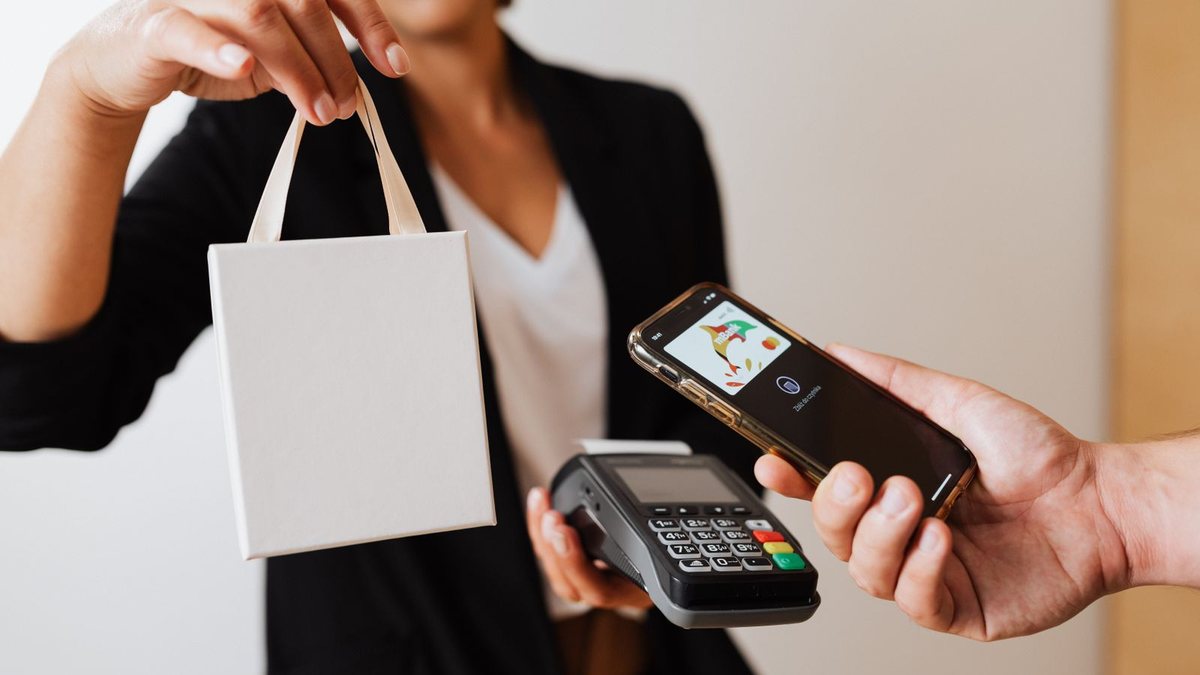 A customer taps a phone against a card terminal while receiving a shopping bag, illustrating a purchase consumed outside the app