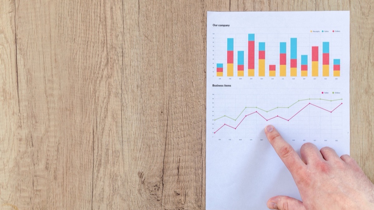 A finger pointing at a rising sales-and-orders trend line on a printed monthly report on a wooden desk