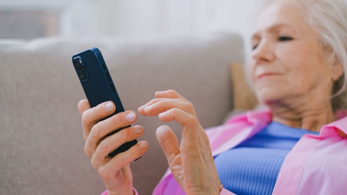 An older woman in a pink shirt sitting on a couch, holding and looking at a smartphone