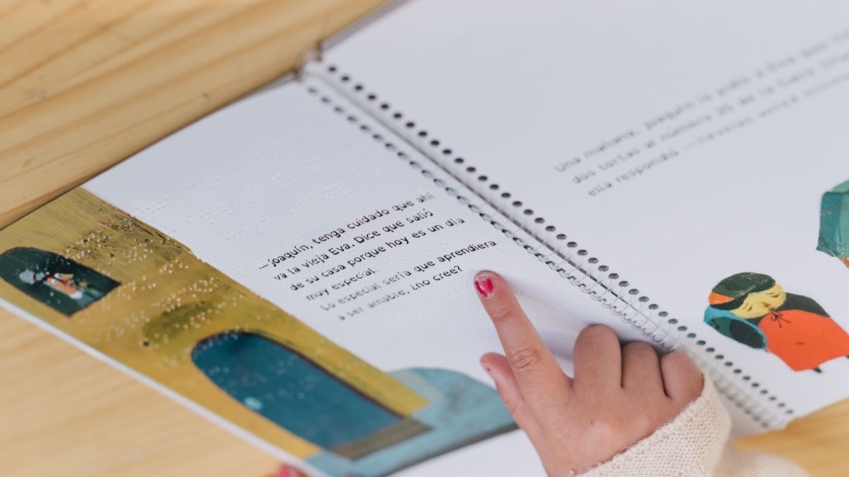 A child's finger resting on a page of an open tactile picture book that combines printed text, colour illustrations and raised Braille dots