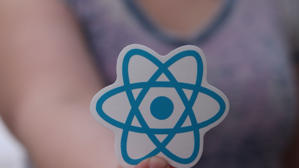 A person holding up a React logo sticker between two fingers against a blurred background