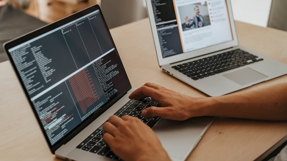 Two laptops side by side on a wooden desk with a developer typing code on one of them