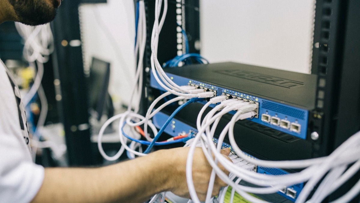 A technician in a white t-shirt plugging white network cables into a blue switch in an equipment rack